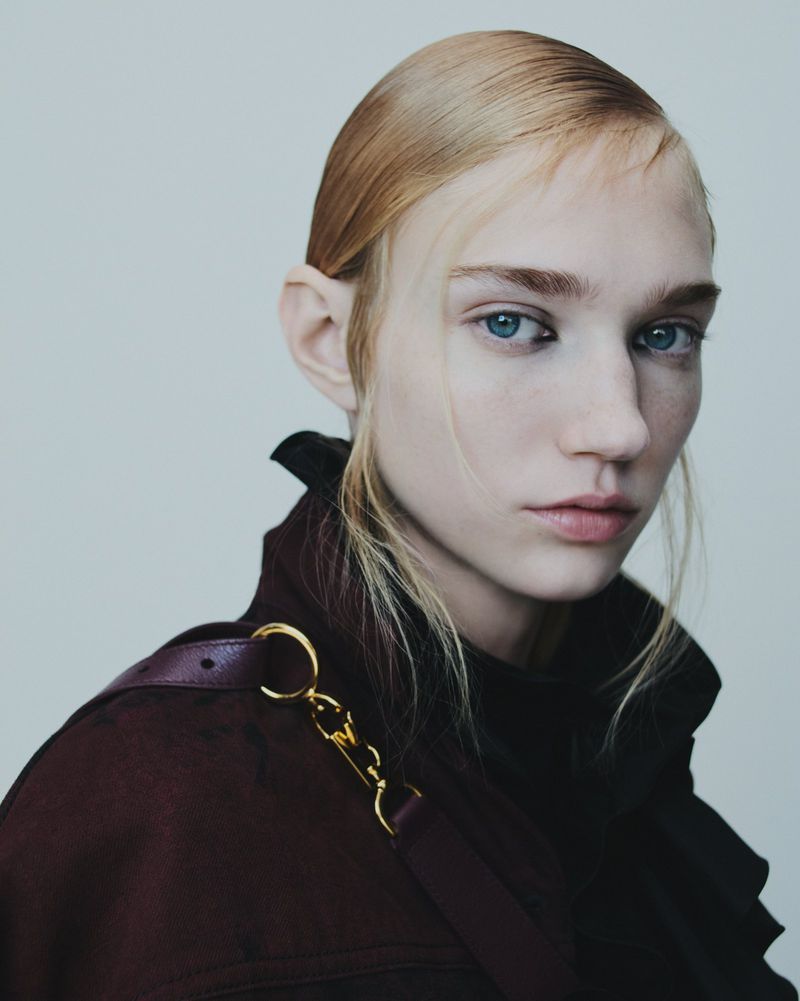 Close-up portrait of a model with sleek hair and blue eyes wearing a dark burgundy coat against a neutral grey studio background.