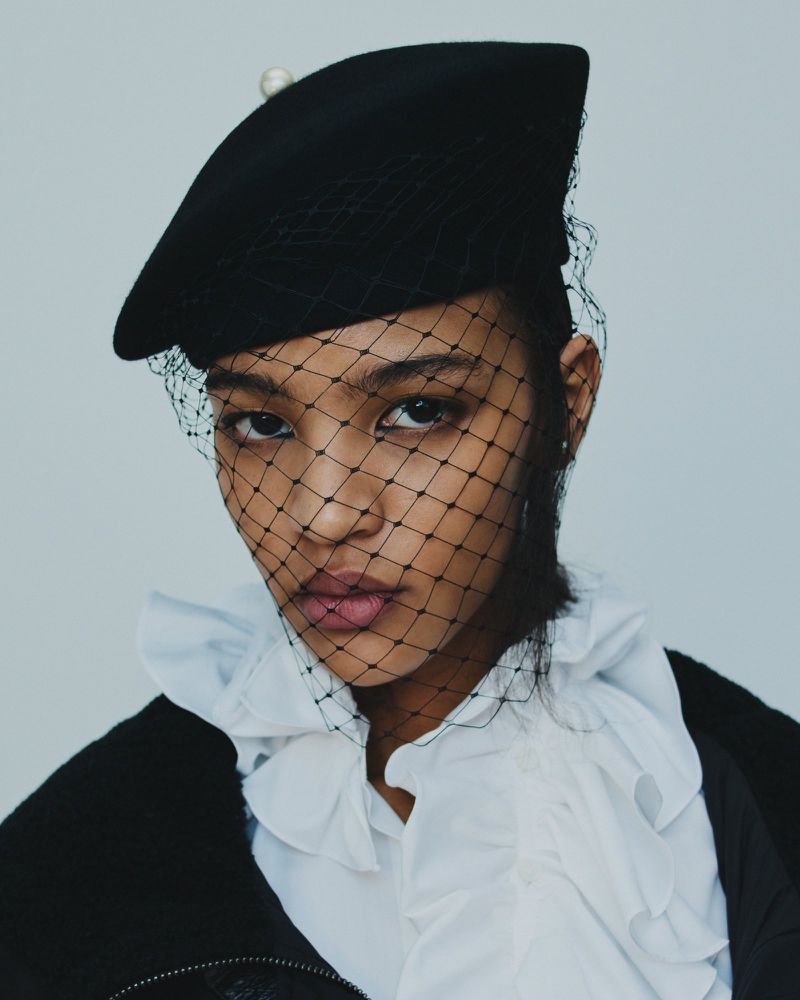 Portrait of an adult female model with fair skin wearing a black beret with netting, a white ruffled collar, and a black jacket.