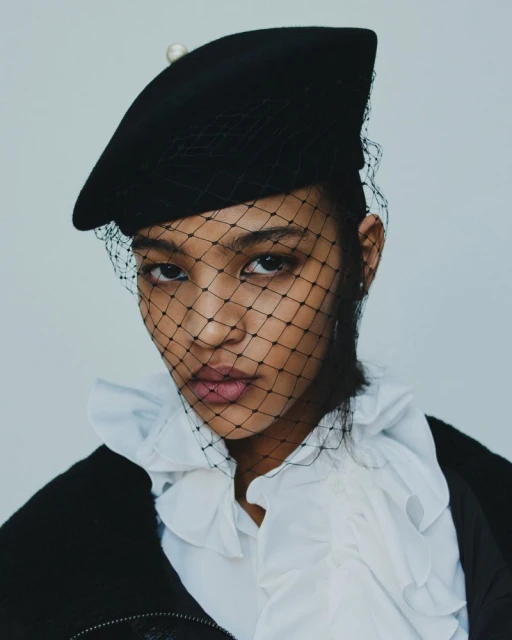 Portrait of an adult female model with fair skin wearing a black beret with netting, a white ruffled collar, and a black jacket.