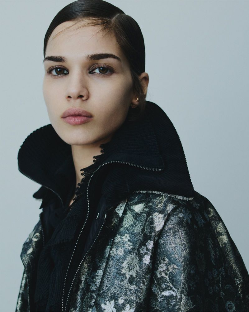 Close-up portrait of a woman with slicked-back hair, wearing a patterned jacket against a neutral background, capturing beauty and fashion.