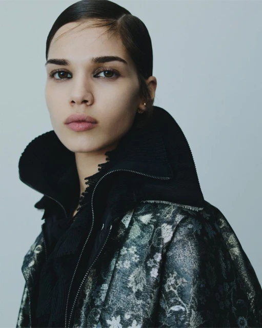 Close-up portrait of a woman with slicked-back hair, wearing a patterned jacket against a neutral background, capturing beauty and fashion.