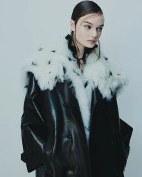 Model wearing a black leather coat with a white faux fur collar against a seamless grey studio background.