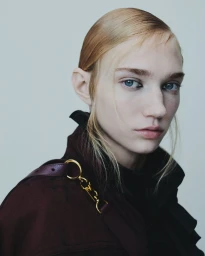 Close-up portrait of a model with sleek hair and blue eyes wearing a dark burgundy coat against a neutral grey studio background.