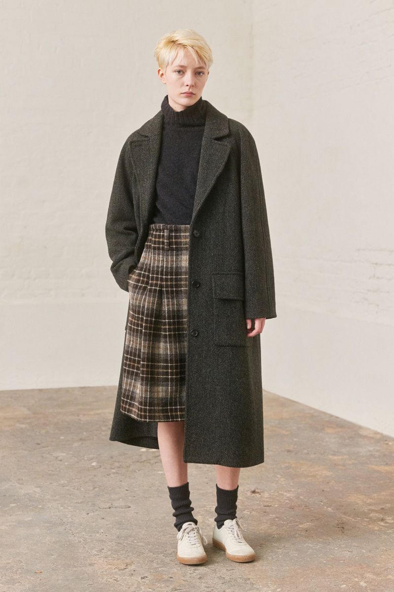 Model with blonde hair wearing dark grey wool coat, plaid skirt, turtleneck, and white sneakers against neutral studio backdrop.