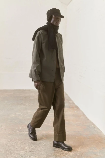 Fashion shot of a male model walking in an olive green shirt jacket, brown pants, a black cap, black scarf, and black leather shoes.