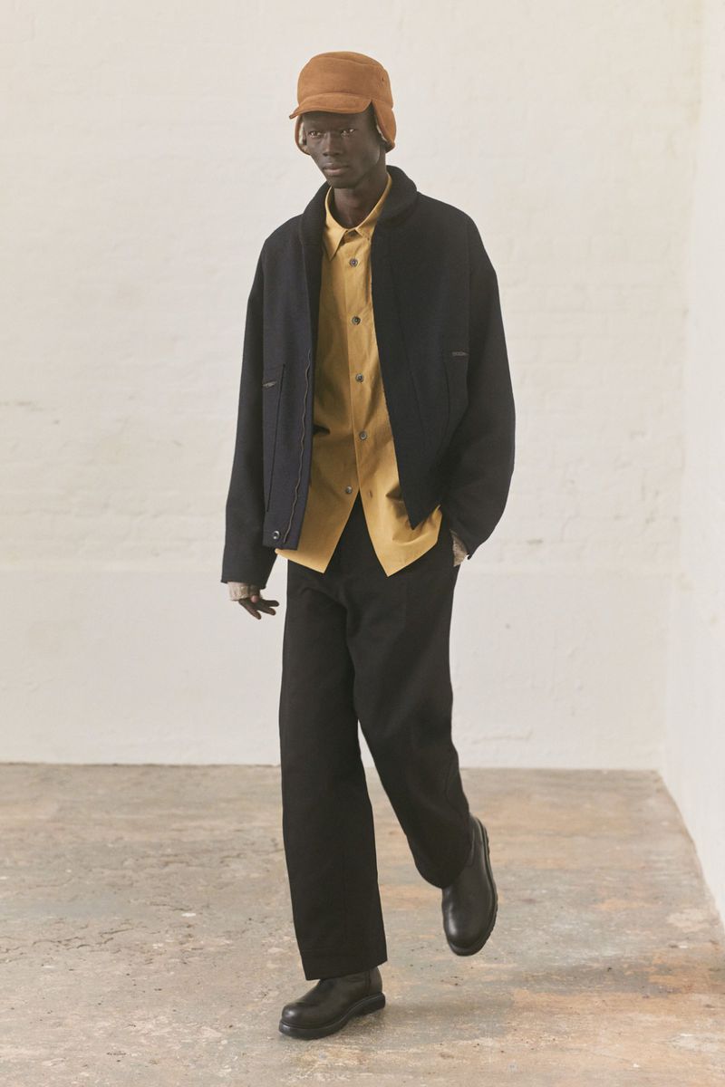 Adult man wearing a tan cap, yellow button-down shirt, and black jacket and pants, posing in a light-filled indoor photo studio.