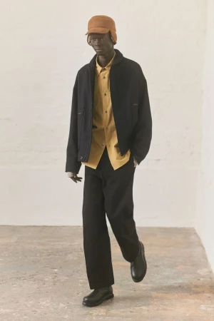 Adult man wearing a tan cap, yellow button-down shirt, and black jacket and pants, posing in a light-filled indoor photo studio.