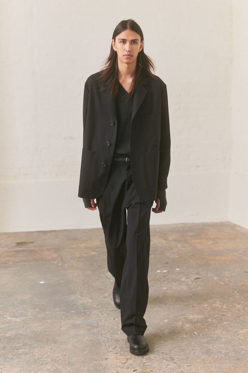 Model with long dark hair wearing a black jacket and trousers walking forward on a weathered stone floor.