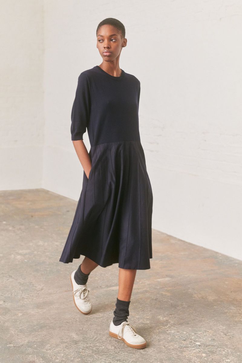 A woman models a midi-length, A-line, navy dress with short sleeves and cream-colored sneakers in a minimally decorated studio setting.