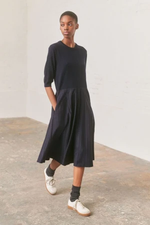 A woman models a midi-length, A-line, navy dress with short sleeves and cream-colored sneakers in a minimally decorated studio setting.