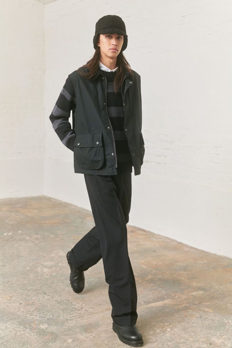 Male model showcases black jacket, striped sweater, and wide-leg trousers indoors, walking toward camera, neutral background, fashion forward.