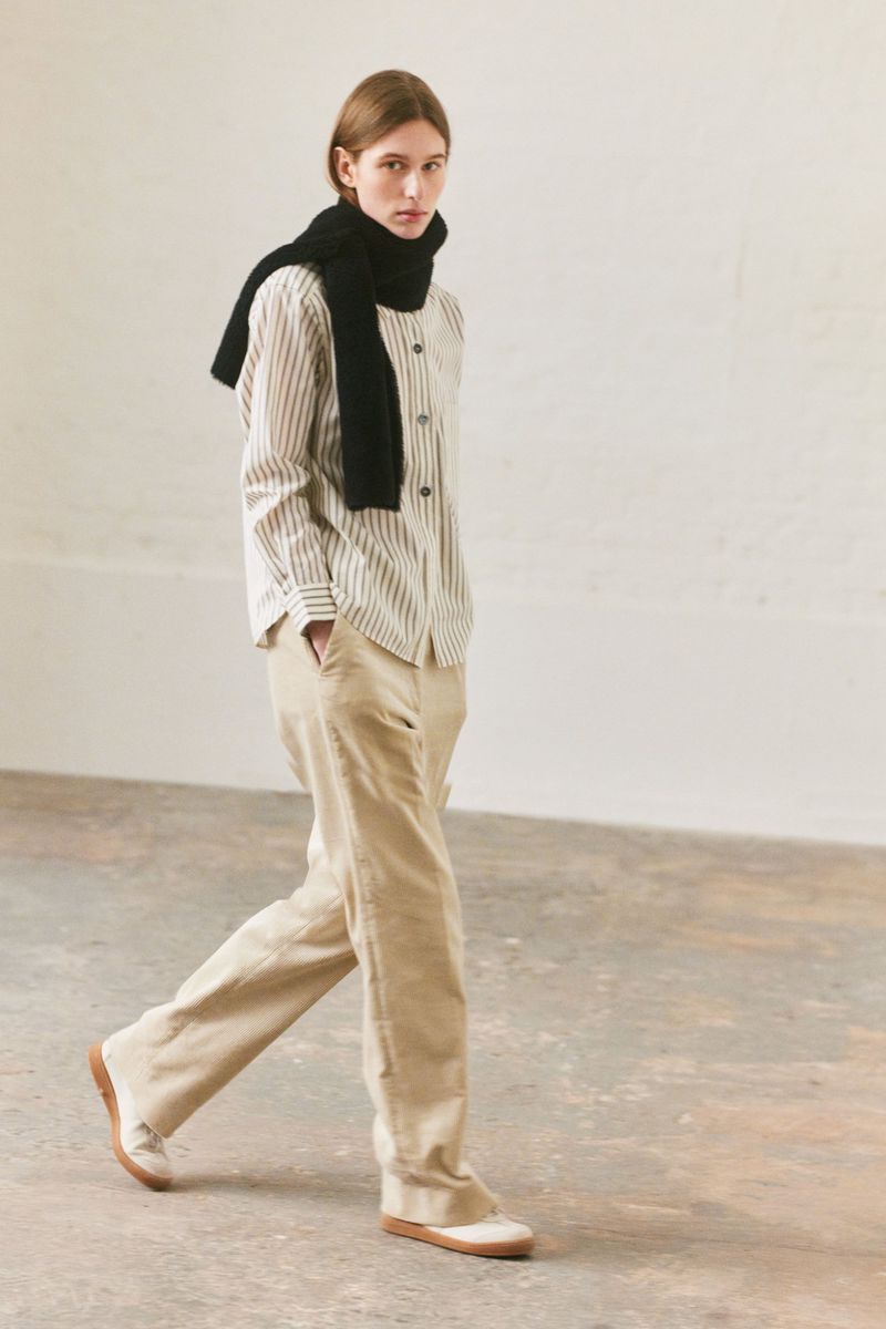 Model walking in a striped shirt, beige trousers, and a black scarf for Margaret Howell F/W 25.