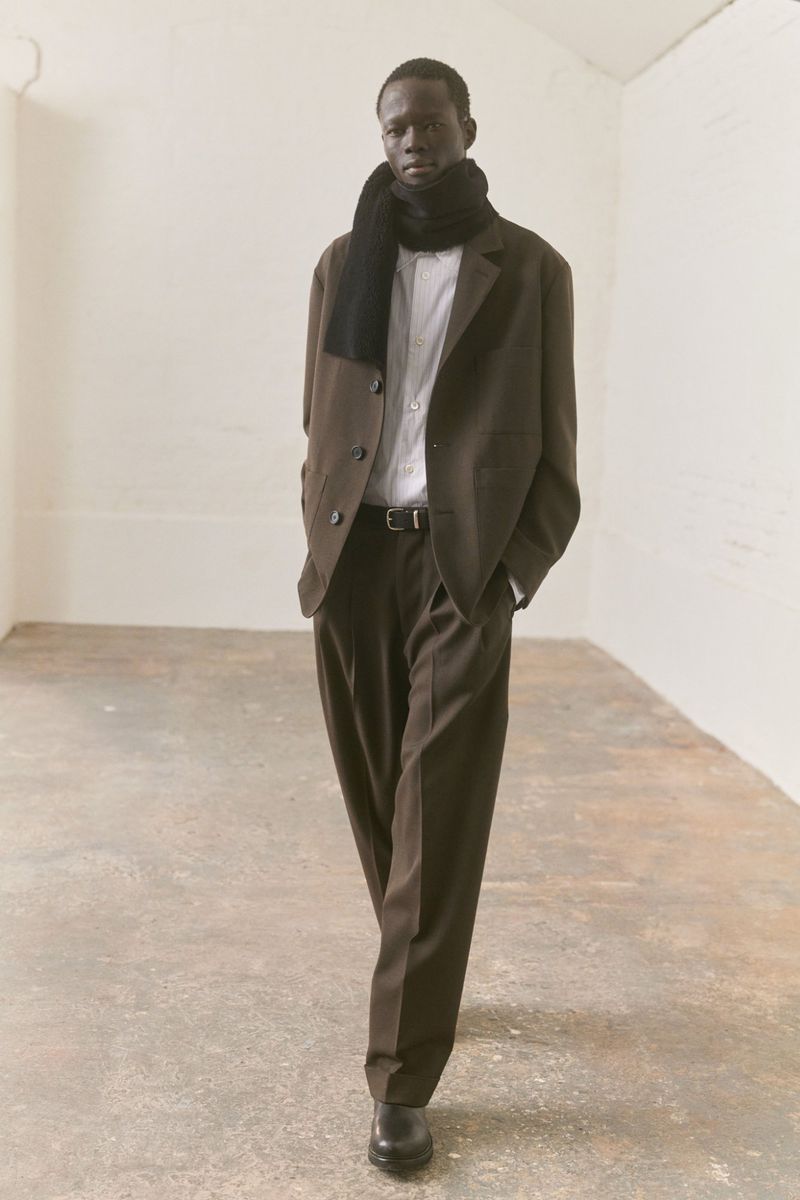 A slender Black male model confidently strides in a brown suit and black scarf against a pale interior backdrop, showcasing fashion.