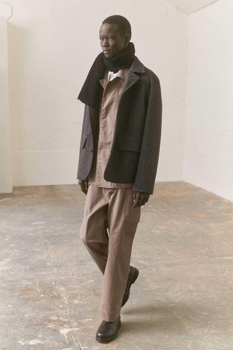 A dark-skinned man in a grey jacket, tan shirt and pants, and a dark scarf walks on a light grey concrete floor against a cream wall.