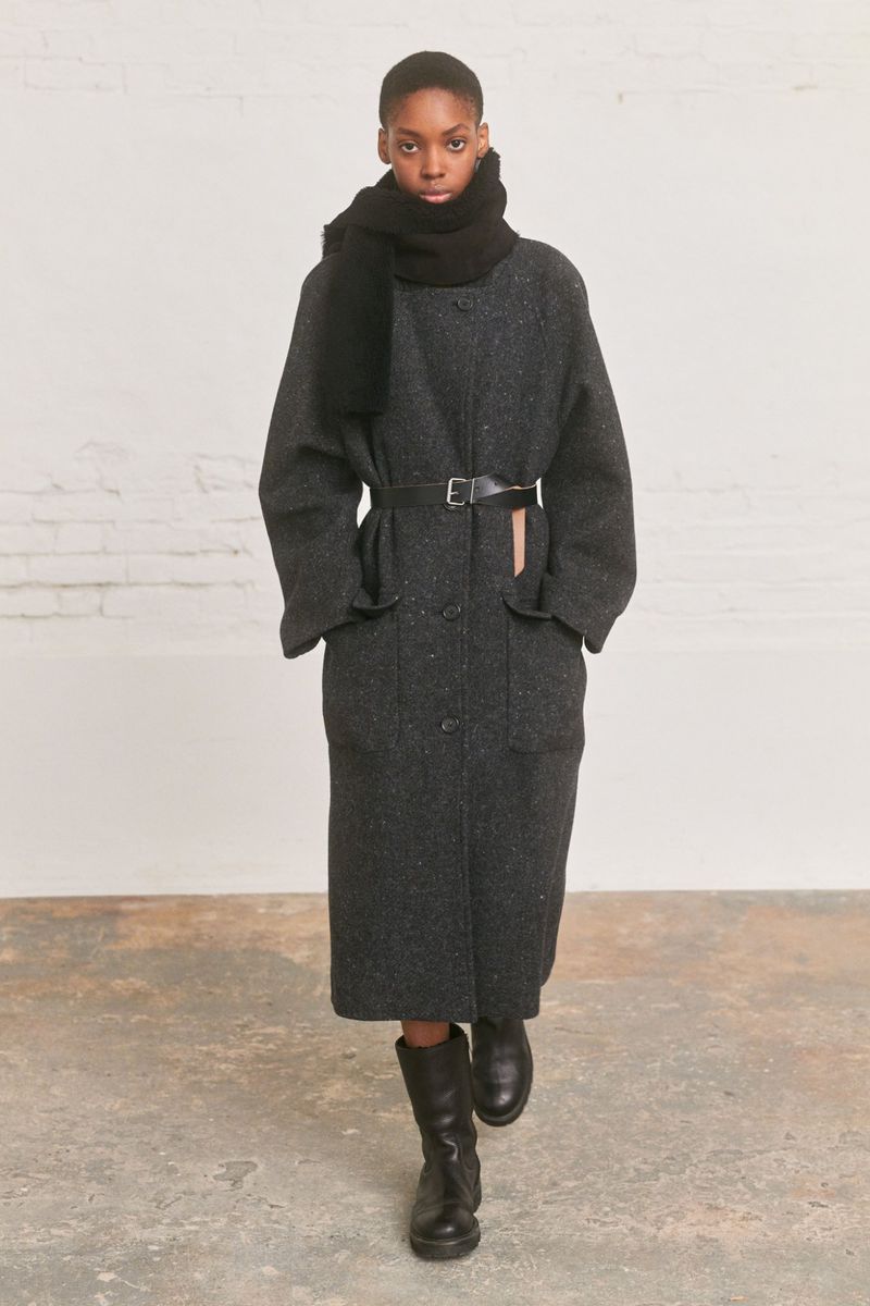 Runway shot of a Black female fashion model wearing a long, speckled grey belted coat, black scarf, and black boots against a plain background.