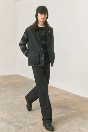 Model in black vest, striped sweater, and bucket hat walking against a neutral textured studio background.