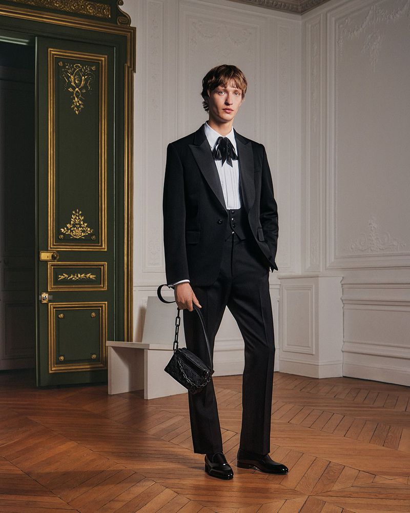 A stylish young adult man in a black tuxedo with bow tie, holding a small black purse, stands in an elegant room with parquet flooring.