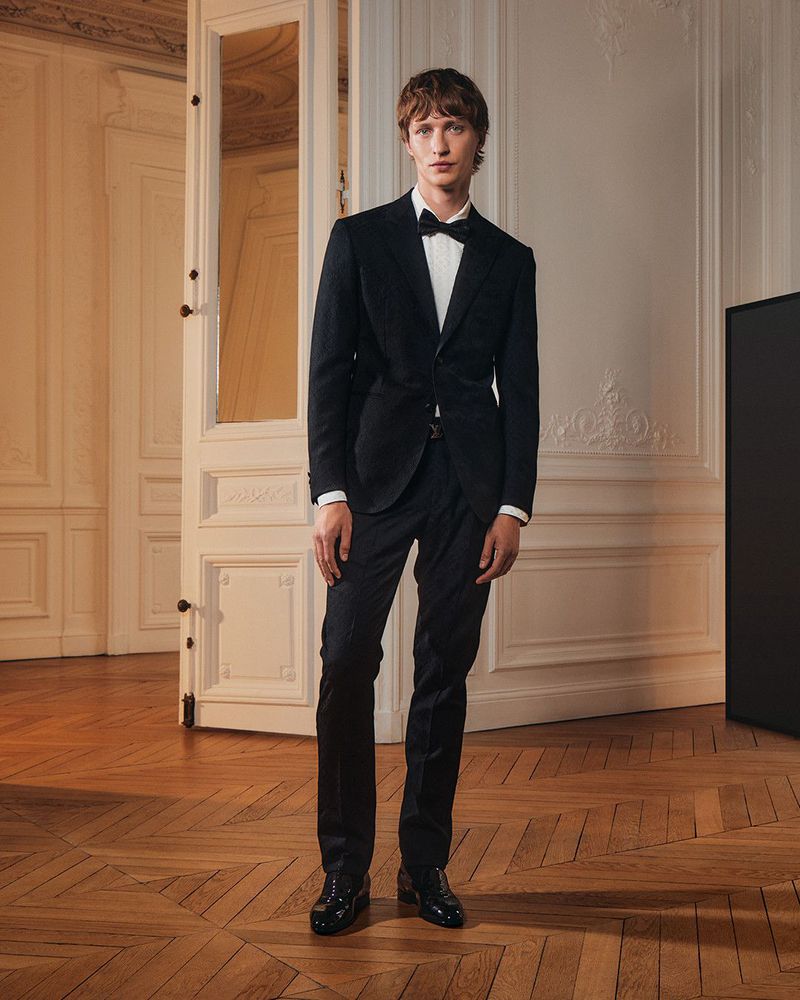 Full-length shot of a man in a black suit with a bow tie standing indoors with wood flooring and white walls and doors.