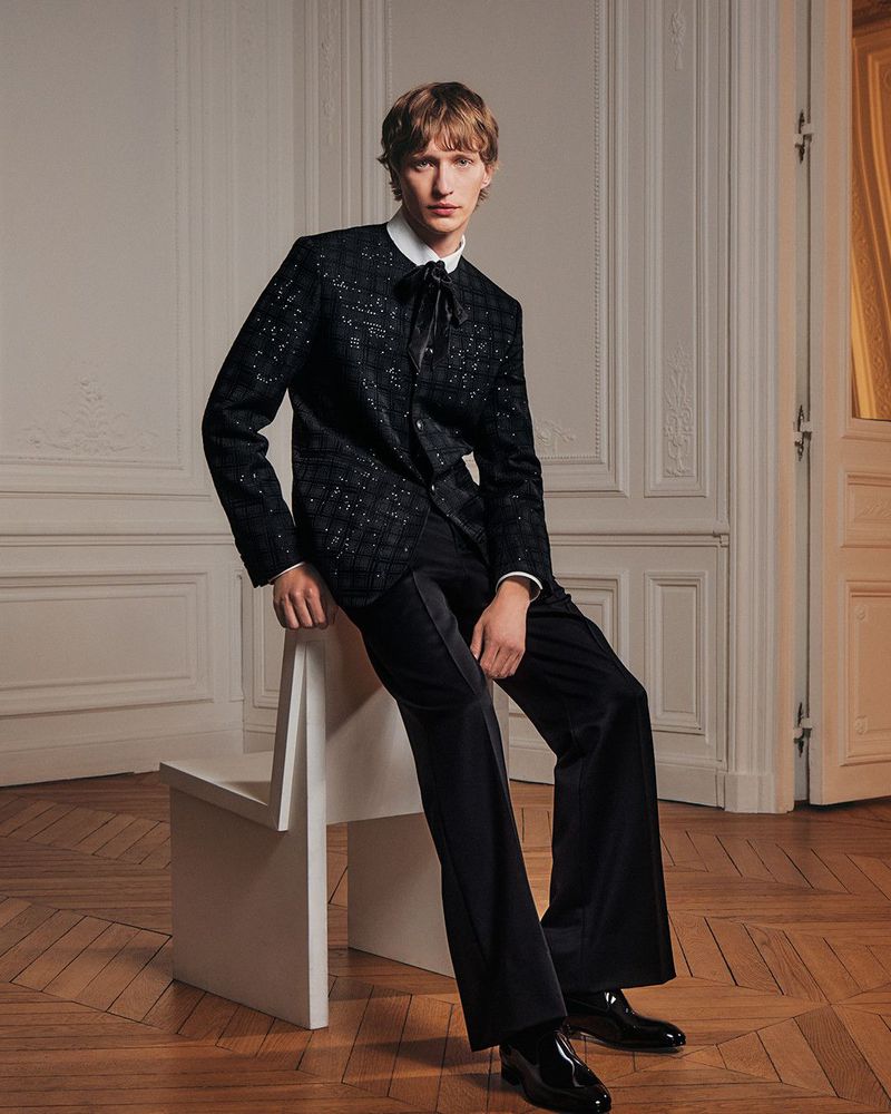 Model seated on a white angular chair wearing a black textured jacket and trousers against ornate panelled walls.