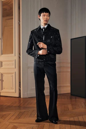 Asian male fashion model in a black embellished jacket and black pants standing indoors in a parquet-floored room with doors and a black screen.