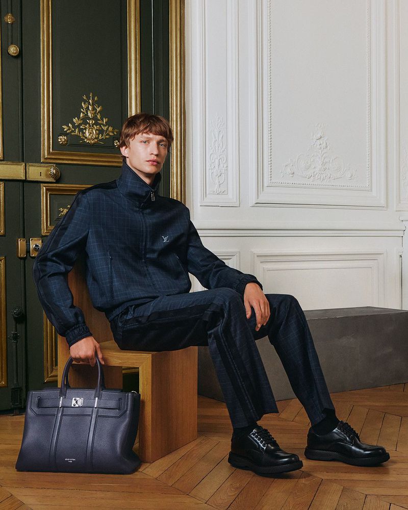 A male fashion model sits on a wooden block with a black leather handbag at his side, wearing a blue tracksuit and black dress shoes.