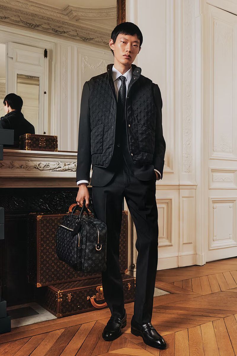 Asian man models a quilted vest and suit while holding a handbag in an elegant interior with Louis Vuitton luggage.