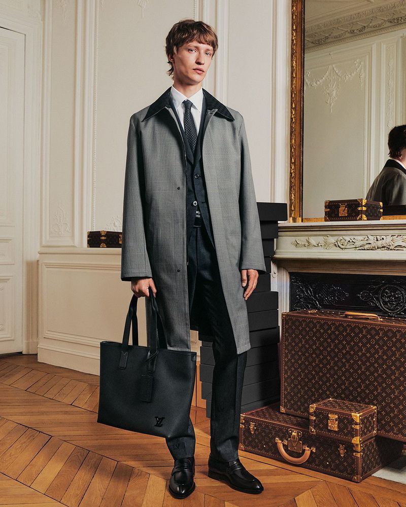 Man in a grey coat and black suit holding a black tote bag, indoors with luggage and decorative fireplace in the background.