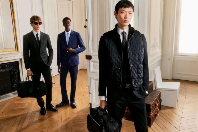 Three men in suits stand inside an elegant room with wood floors and vintage luggage; the closest holds a Louis Vuitton bag.