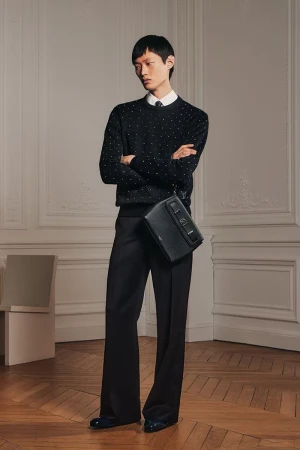 Adult male fashion model poses indoors wearing a dotted sweater, black dress pants, and carrying a black leather messenger bag.