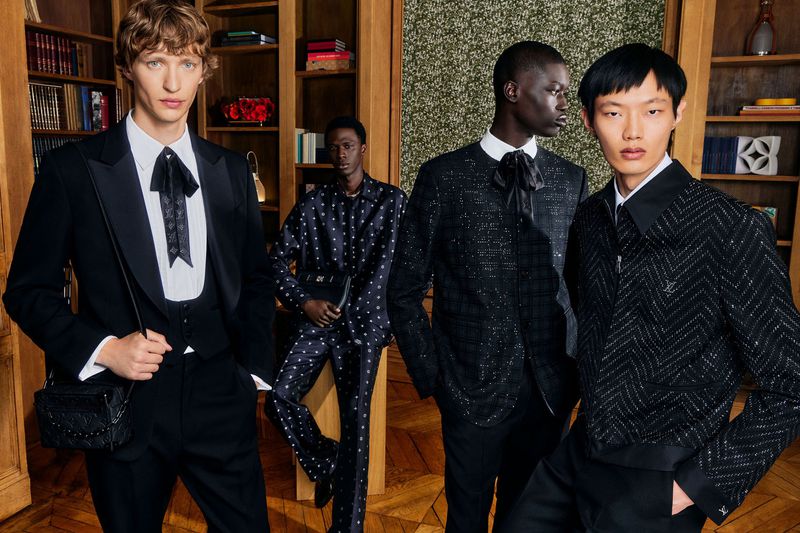 Four young adult men in stylish formalwear pose indoors against a backdrop of dark wood shelving and a mossy feature wall.