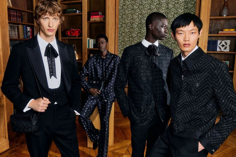 Four young adult men in stylish formalwear pose indoors against a backdrop of dark wood shelving and a mossy feature wall.