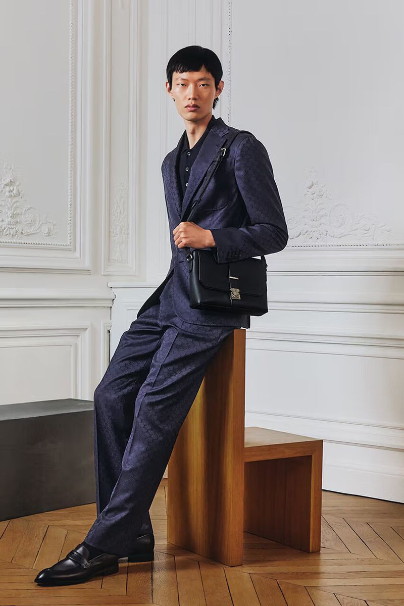 An Asian man in a blue suit poses in a studio with a modern, minimalist aesthetic. He wears a black satchel and black shoes.
