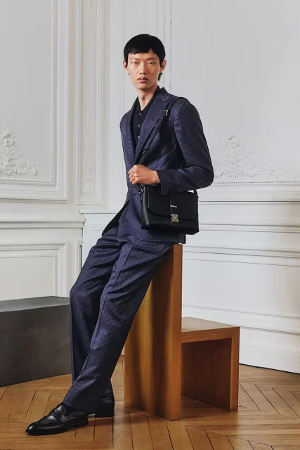 An Asian man in a blue suit poses in a studio with a modern, minimalist aesthetic. He wears a black satchel and black shoes.