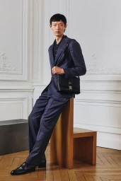 Model wearing a navy suit and black leather shoulder bag sitting on a wooden stool indoors.