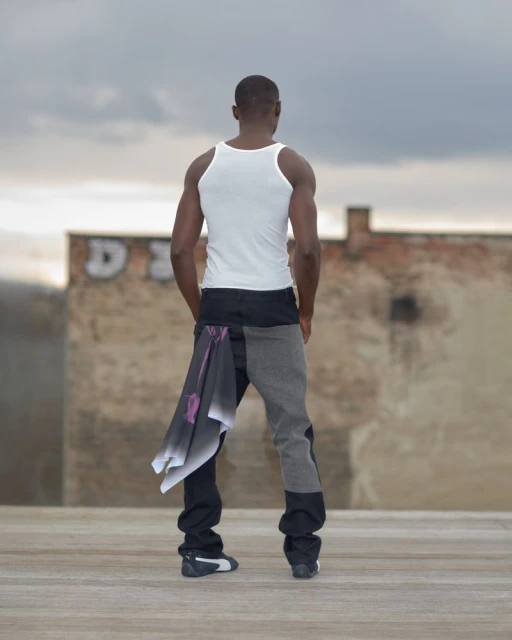 Black man in a white tank top and grey jeans with a scarf, stands on a rooftop, looking at a building against an overcast sky backdrop.