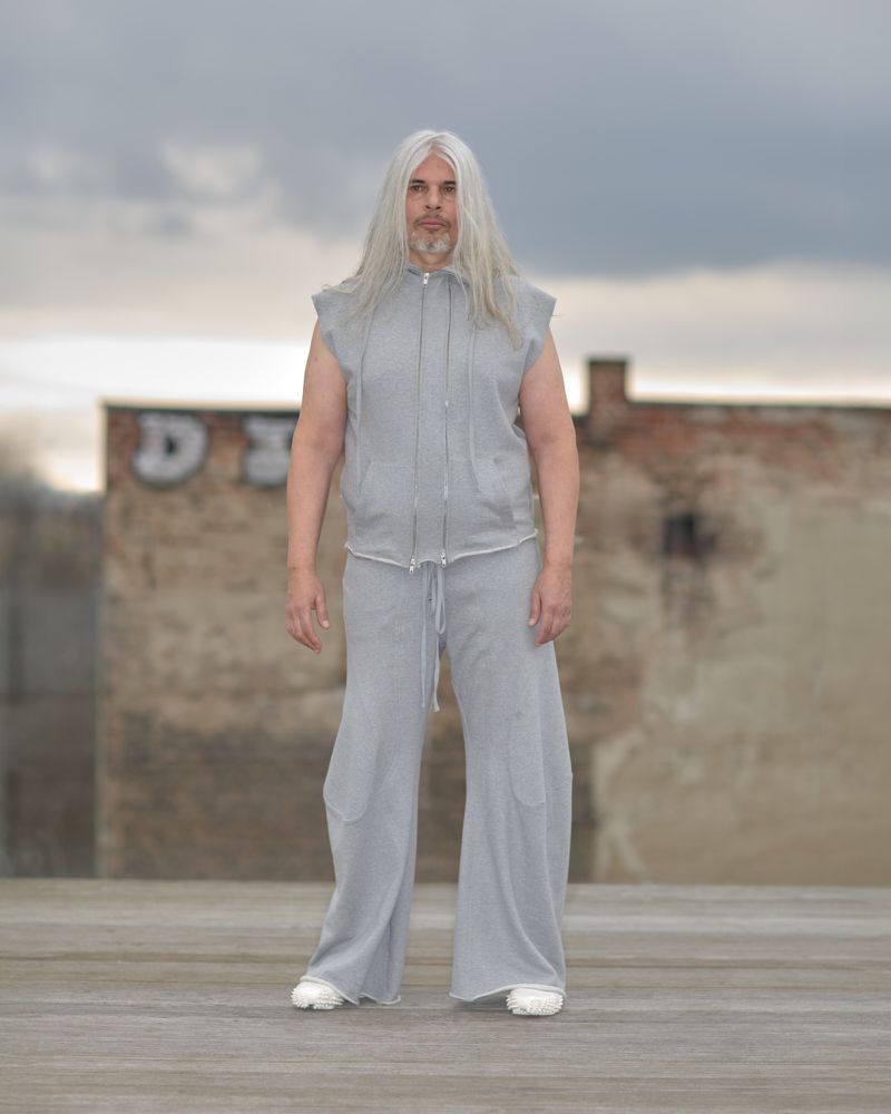 Adult male with long gray hair stands on an outdoor rooftop wearing a gray sleeveless hoodie and matching wide-leg pants in an urban setting.