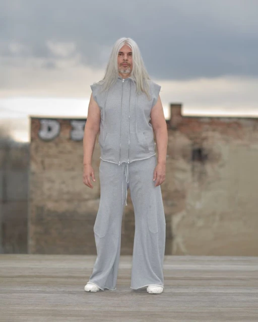 Adult male with long gray hair stands on an outdoor rooftop wearing a gray sleeveless hoodie and matching wide-leg pants in an urban setting.