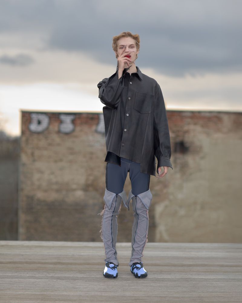 Person in oversized shirt and distressed jeans poses against weathered wall under cloudy sky, fashion-forward and urban-inspired.