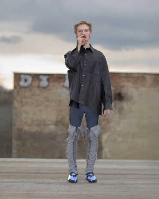 Person in oversized shirt and distressed jeans poses against weathered wall under cloudy sky, fashion-forward and urban-inspired.
