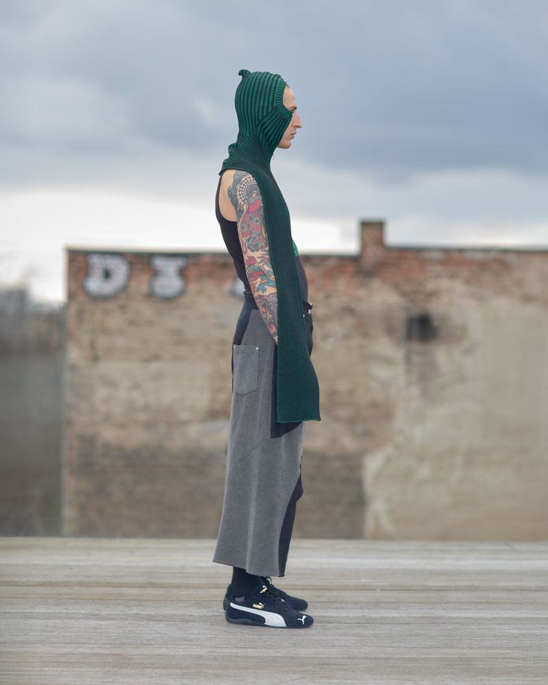 Fashionable person in a green knit balaclava and black tank top stands against an urban backdrop, showcasing streetwear style.
