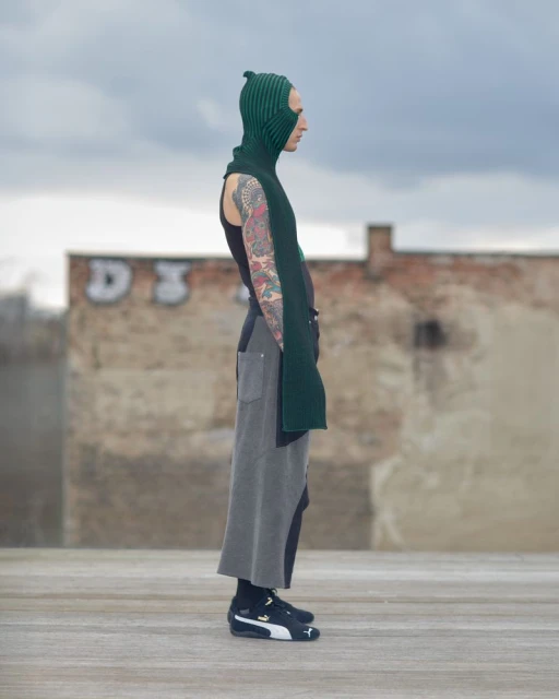 Fashionable person in a green knit balaclava and black tank top stands against an urban backdrop, showcasing streetwear style.
