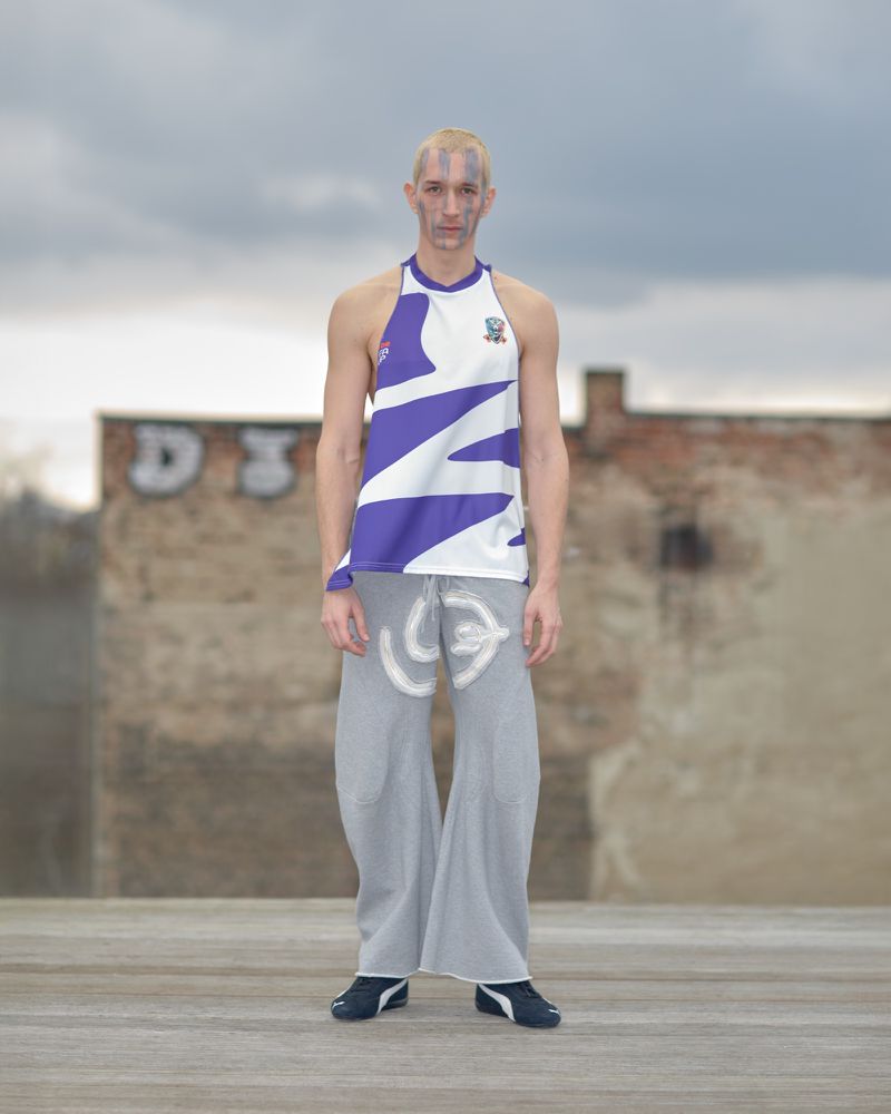 An adult male model poses outdoors wearing a white and purple halter top and gray pants against a backdrop of brick walls and cloudy sky.