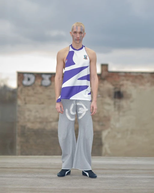 An adult male model poses outdoors wearing a white and purple halter top and gray pants against a backdrop of brick walls and cloudy sky.