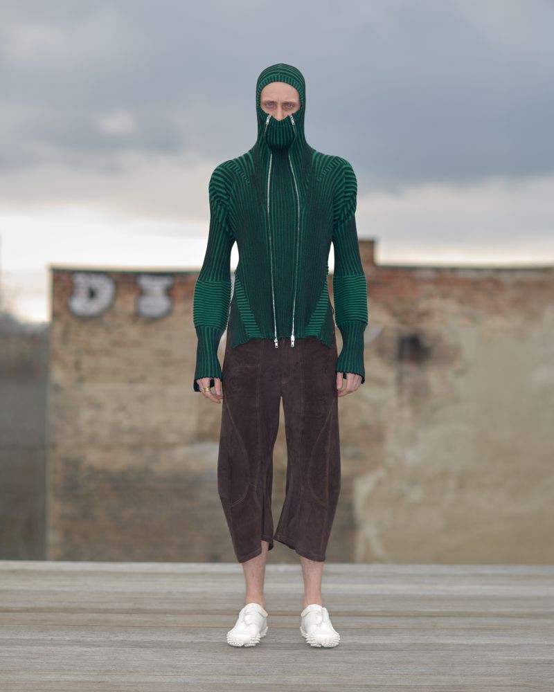 Person wearing a balaclava-style green ribbed sweater with zipper and brown culottes standing outdoors against a cloudy sky and graffiti wall.