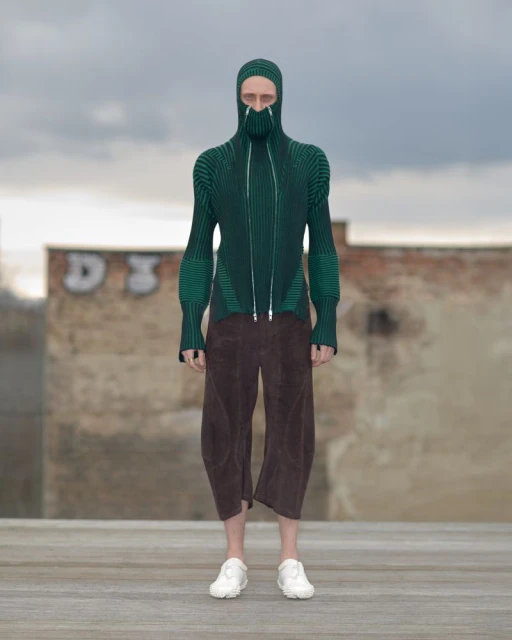 Person wearing a balaclava-style green ribbed sweater with zipper and brown culottes standing outdoors against a cloudy sky and graffiti wall.