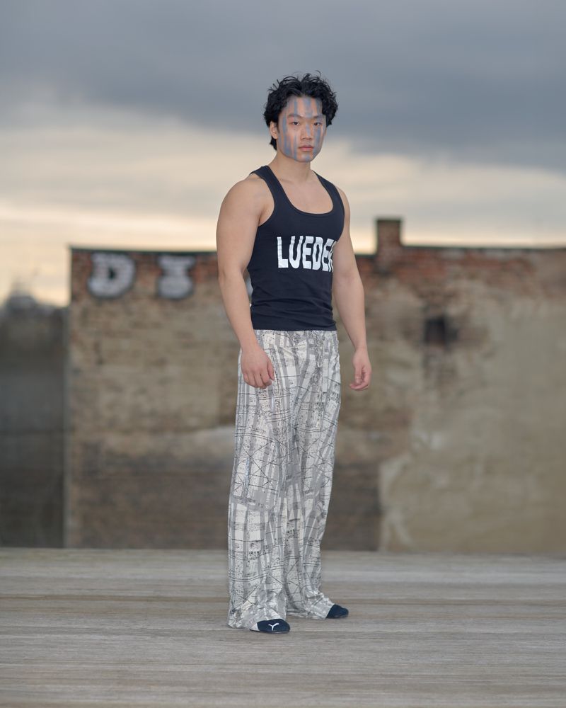 Young Asian male wearing black tank top and patterned pants stands outside against brick wall and cloudy sky. Fashion editorial image.