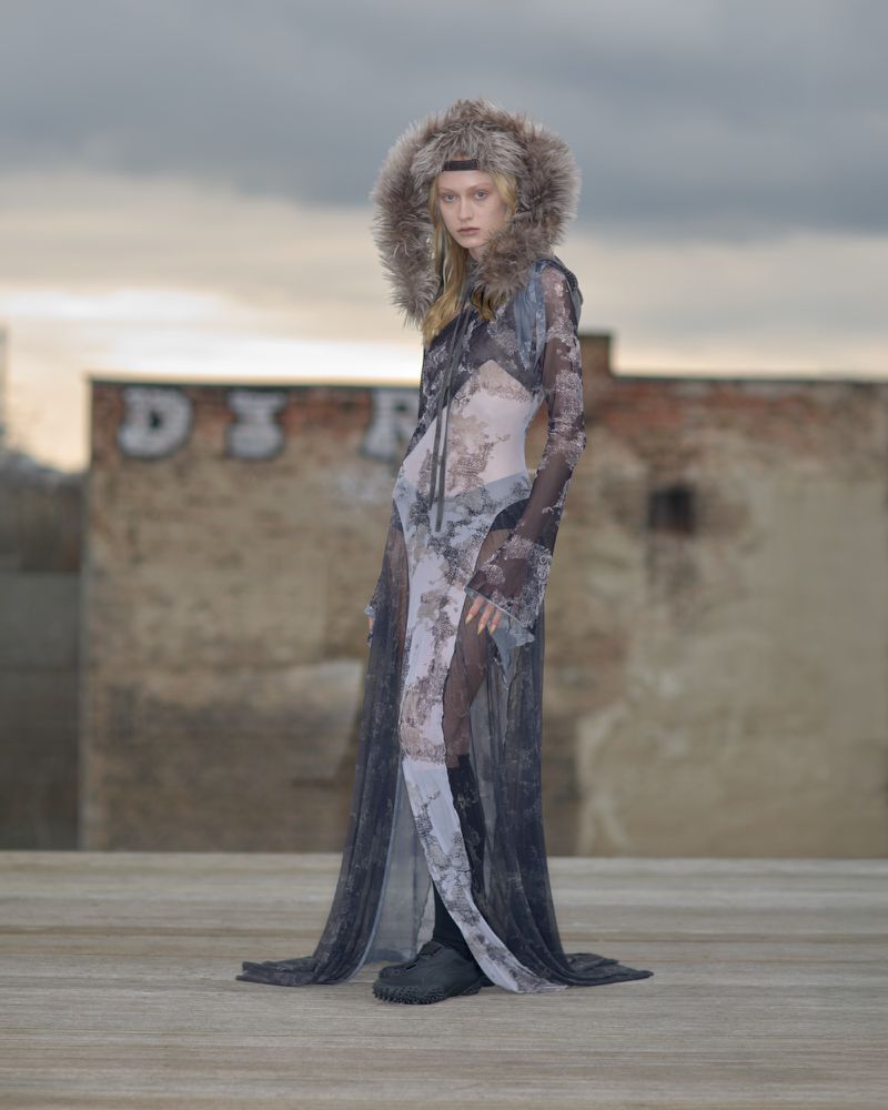 An adult female stands on a flat rooftop wearing a sheer black-and-white patterned dress with a fur hood and black boots against a cloudy sky.
