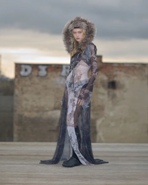 An adult female stands on a flat rooftop wearing a sheer black-and-white patterned dress with a fur hood and black boots against a cloudy sky.