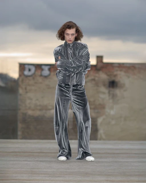 Fashion model poses in a grey velvet jacket and pantsuit with arms crossed outdoors against a cloudy sky and urban background.
