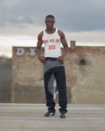 Black man wearing a white printed tank top and dark panelled trousers posing on an outdoor rooftop platform.